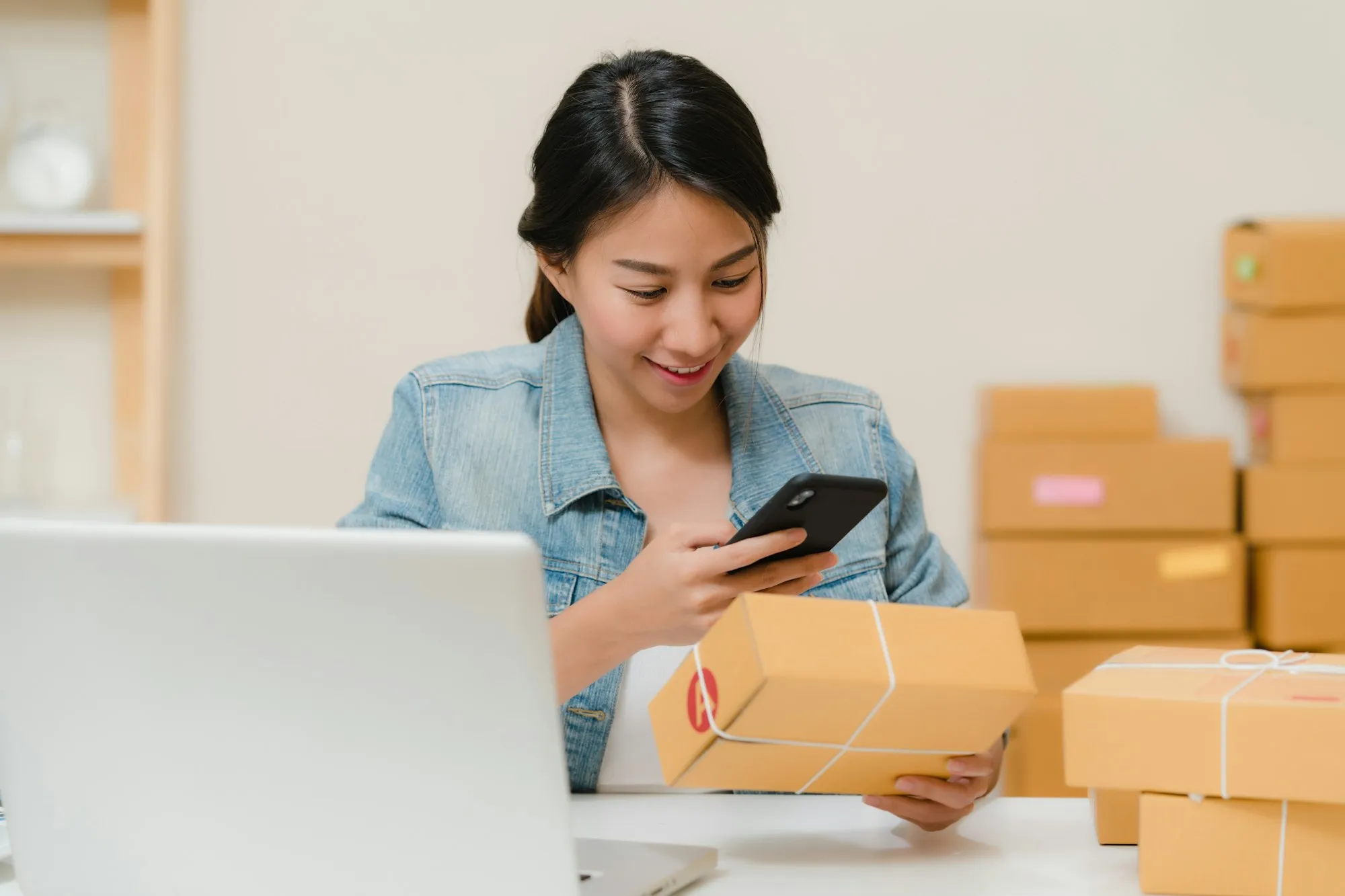 Online store owner checking a parcel against her phone beside a laptop and stacked boxes